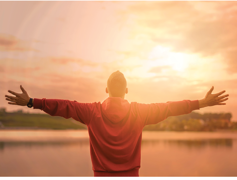 Rear view of man spreading arms and watching the ocean while sun flare is coming trough his arms from behind  Blue ocean and in background and blue sky  Man is wearing red top with hood and black watch on his left wrist